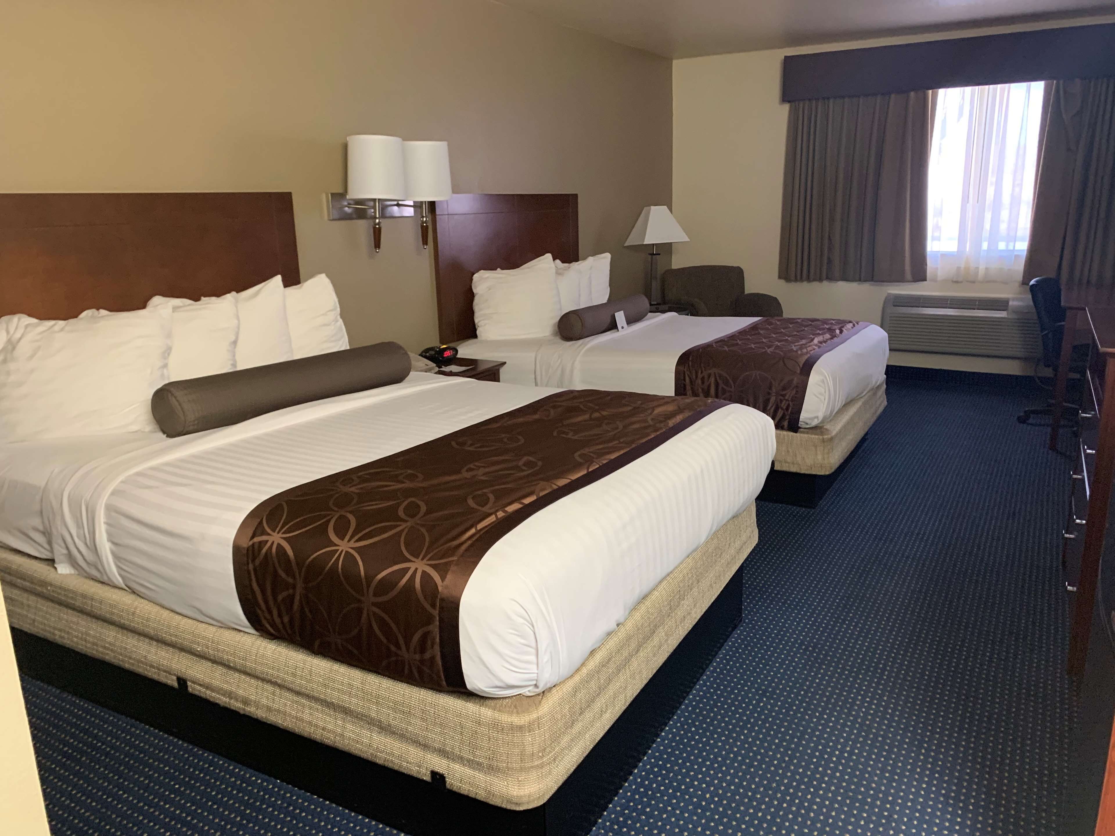A hotel room with two beds, a desk, a chair, a lamp, and a window with curtains.