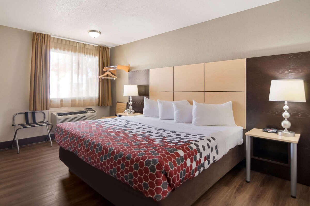 A hotel room with a large bed, a bedside table with a lamp, a chair, and a window with curtains.