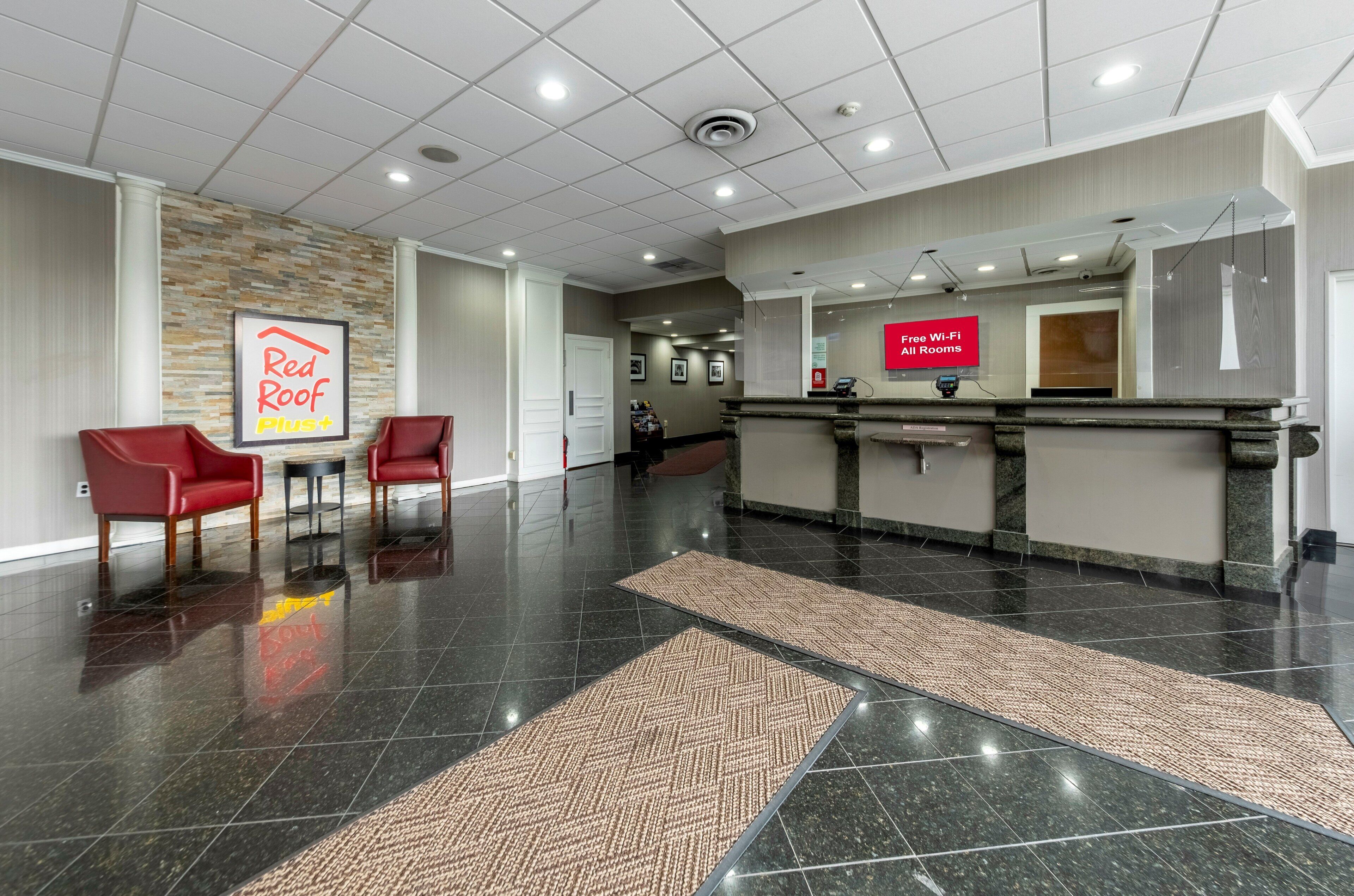 A hotel lobby with a reception desk, two red chairs, a Red Roof Plus sign, and a sign offering free Wi-Fi in all rooms.