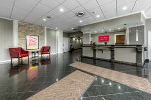 A hotel lobby with a reception desk, two red chairs, a Red Roof Plus sign, and a sign offering free Wi-Fi in all rooms.