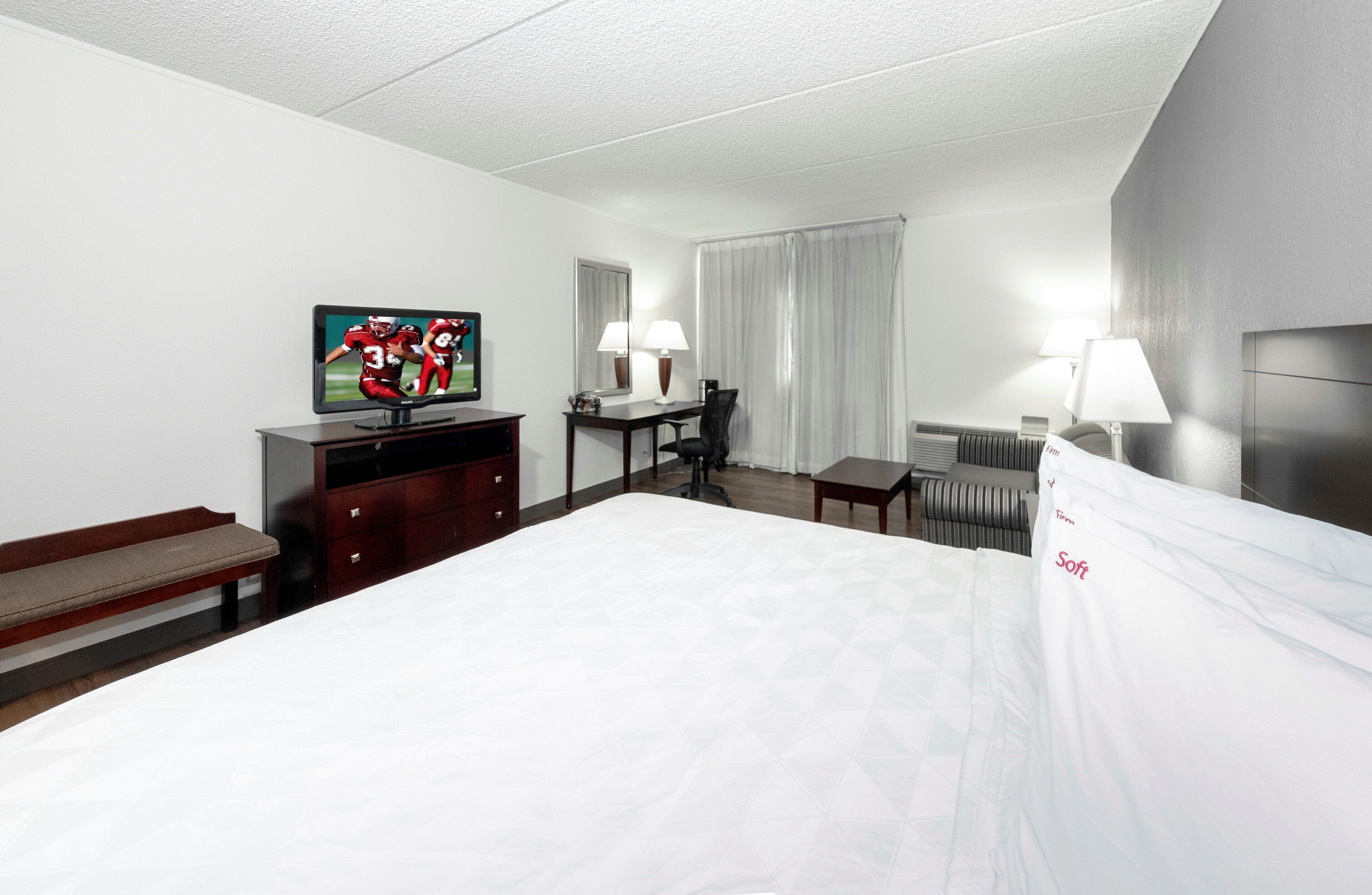 Une chambre d’hôtel avec un grand lit, un bureau avec une chaise, une télévision, une lampe et une fenêtre avec des rideaux.