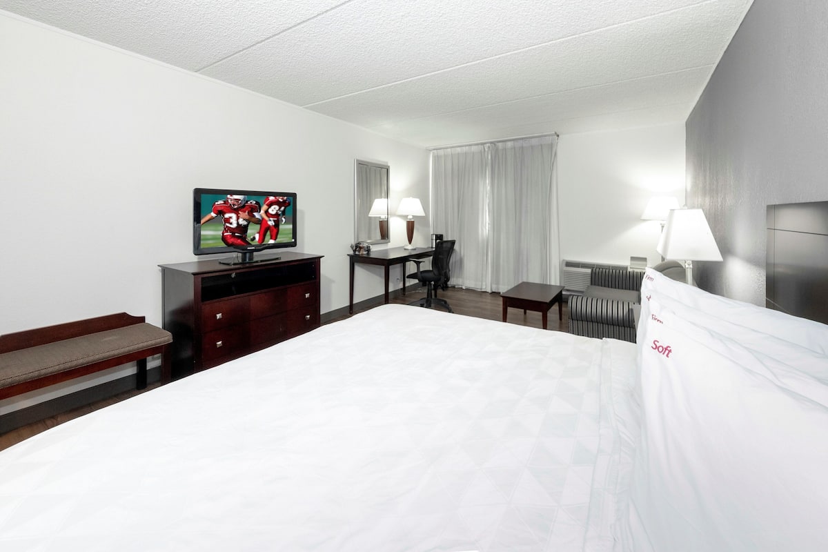 A hotel room with a large bed, a desk with a chair, a television, a bench, and a window with curtains.