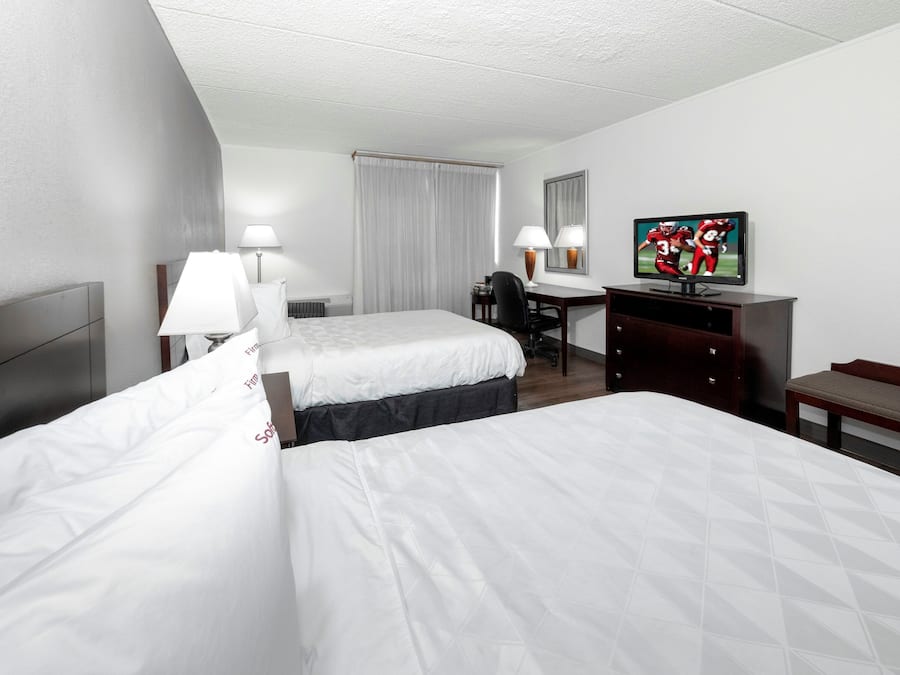 A hotel room with two beds, a desk, a television, and a bench.