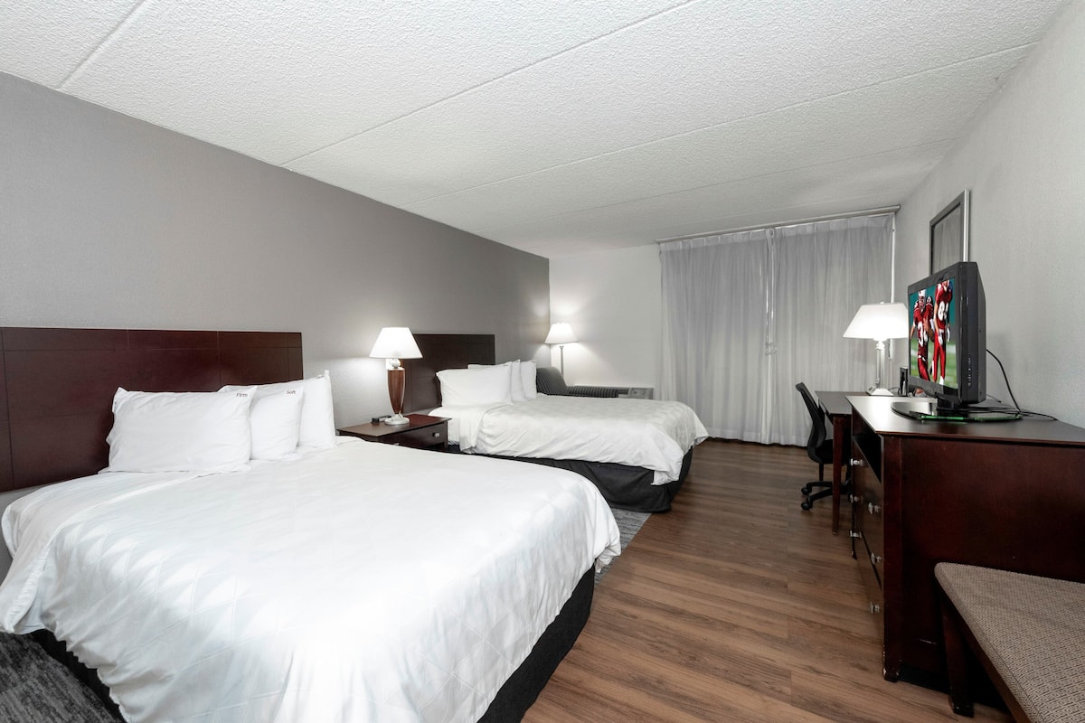A hotel room with two beds, a desk, a chair, a TV, and a lamp.
