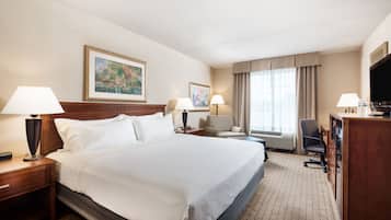A hotel room with a large bed, two bedside tables with lamps, a chair, a desk with a TV, and a large window with curtains.