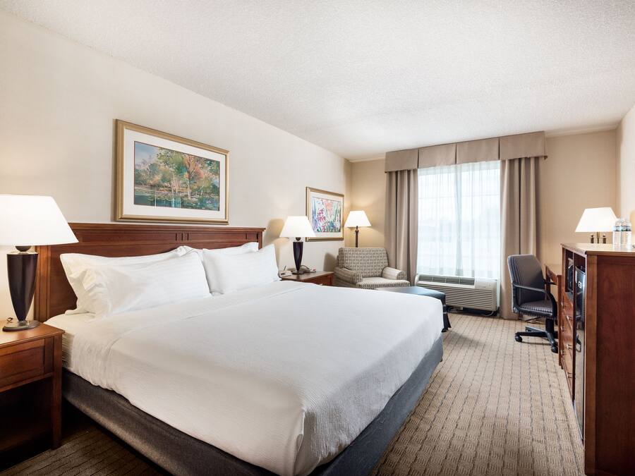 A hotel room with a large bed, two bedside tables with lamps, a chair, a desk with a TV, and a large window with curtains.