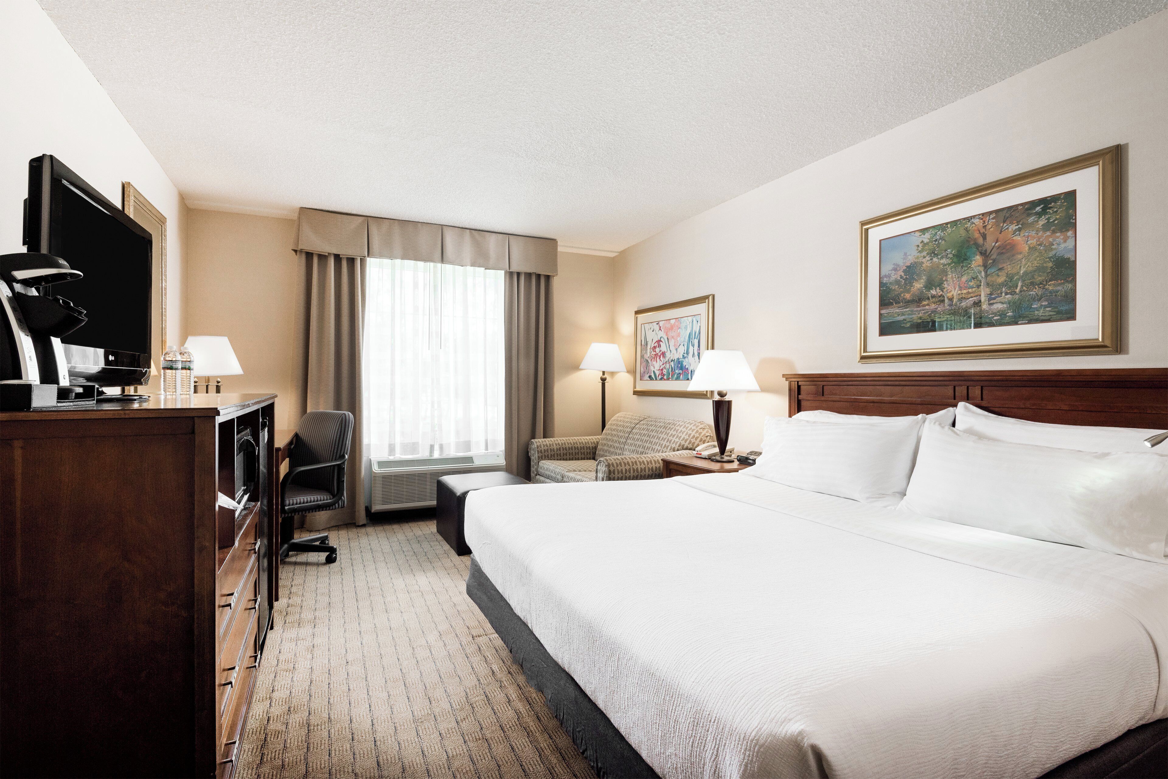 A hotel room with a bed, a desk with a computer, a chair, a window with curtains, a painting on the wall, and a lamp.