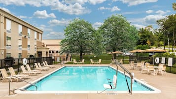 A swimming pool with lounge chairs and umbrellas, surrounded by a fence, with a building in the background.