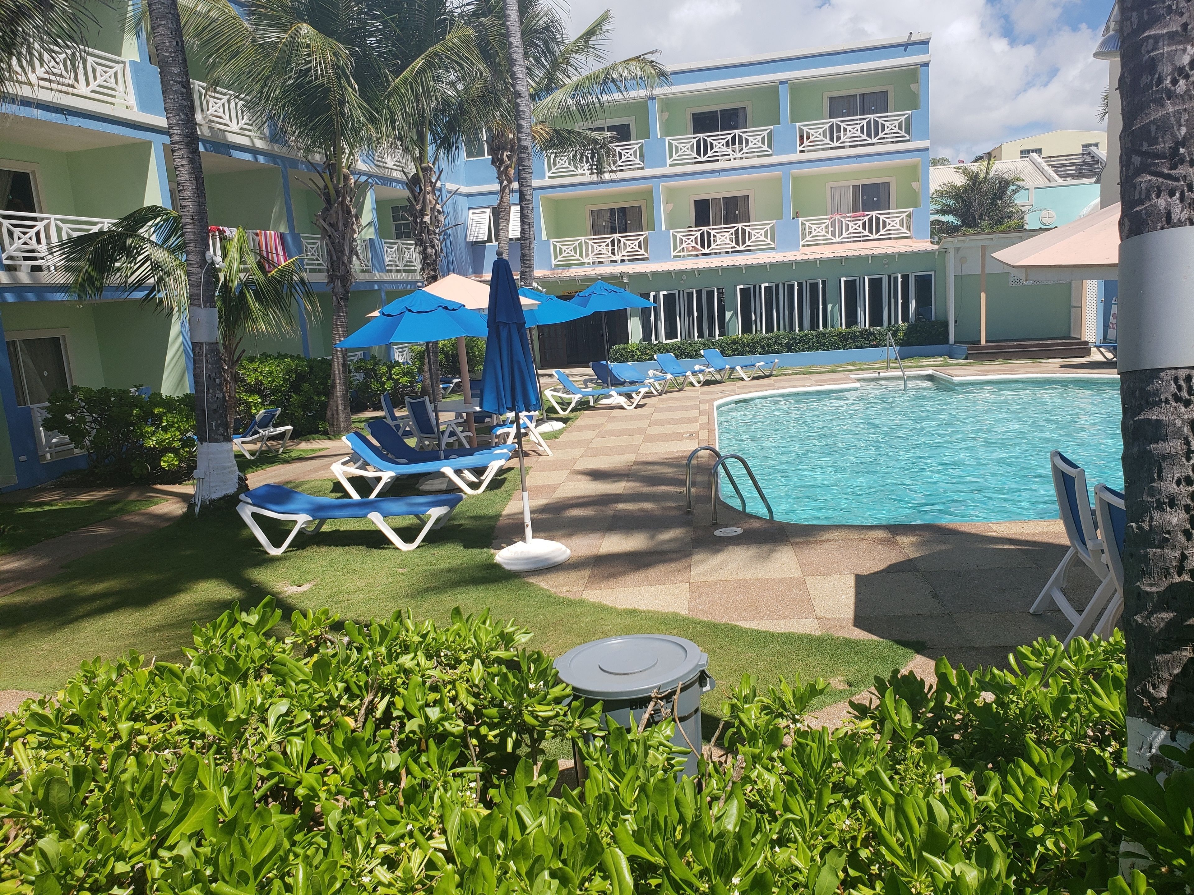 A pool area with lounge chairs and umbrellas, surrounded by greenery and a multi-story building with balconies.