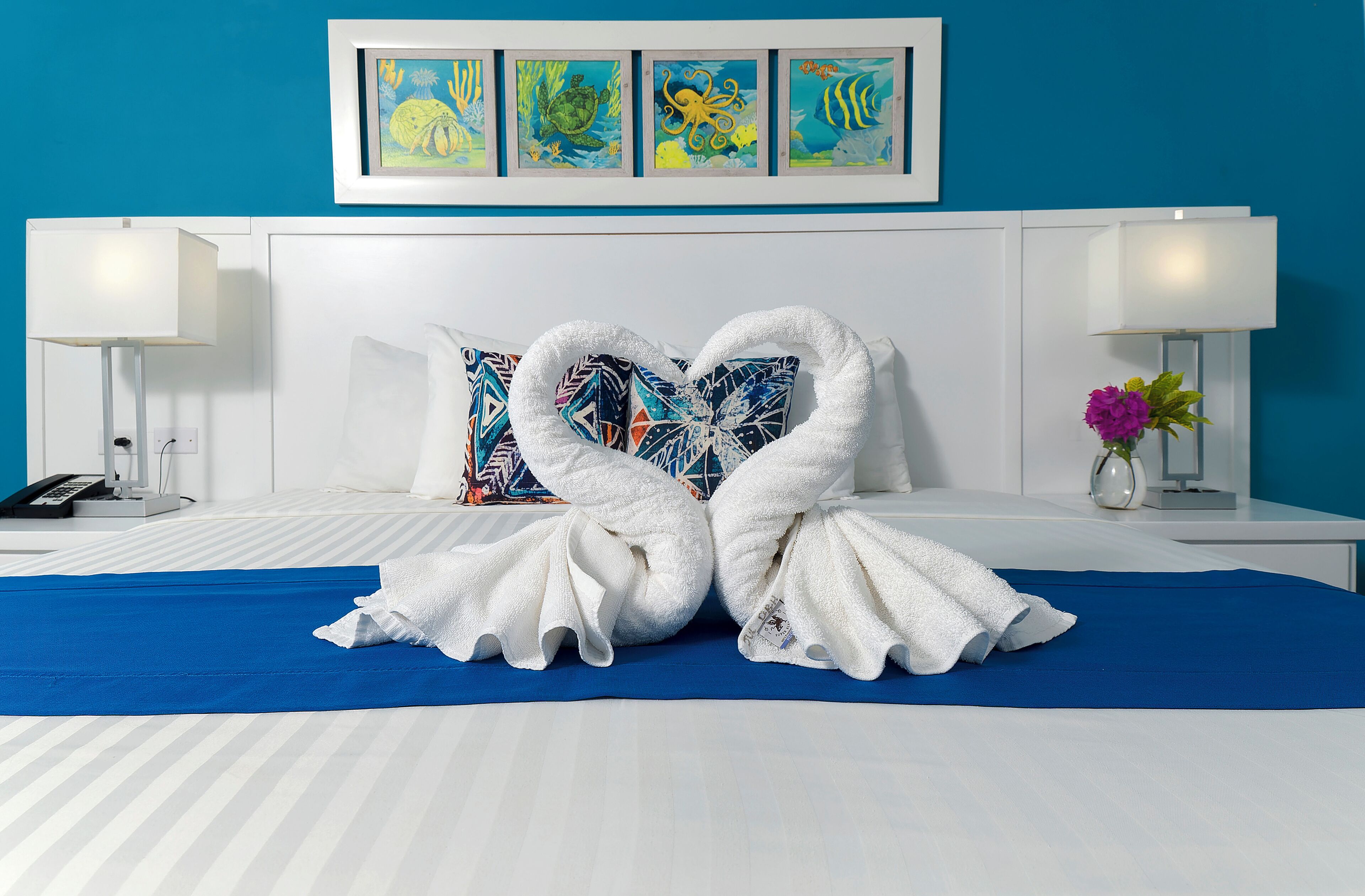A hotel room with a bed, two white towels arranged in a heart shape, a blue bedspread, and a wall with colorful artwork.