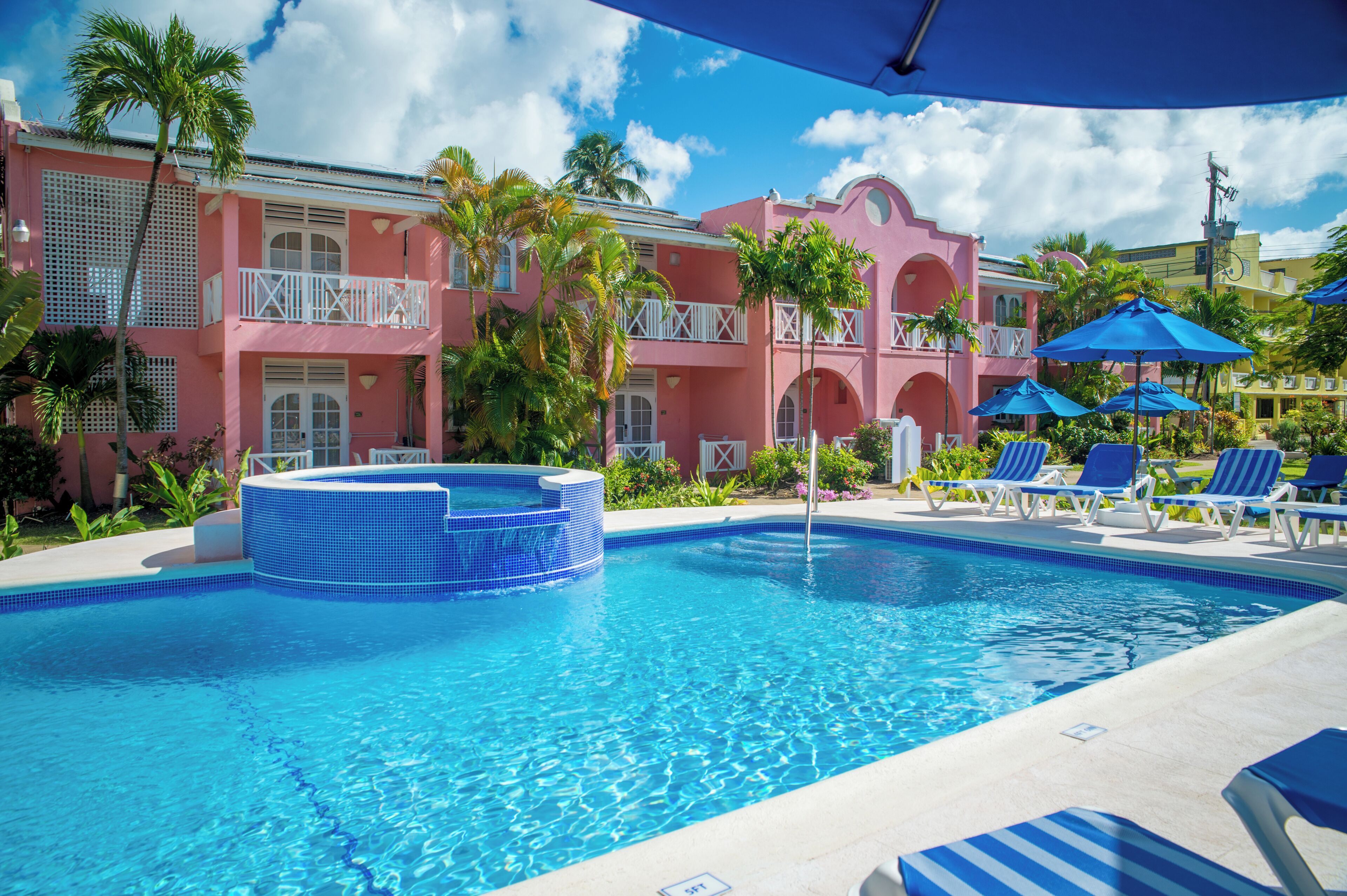 A swimming pool with a hot tub, surrounded by lounge chairs and umbrellas, with a pink building in the background.