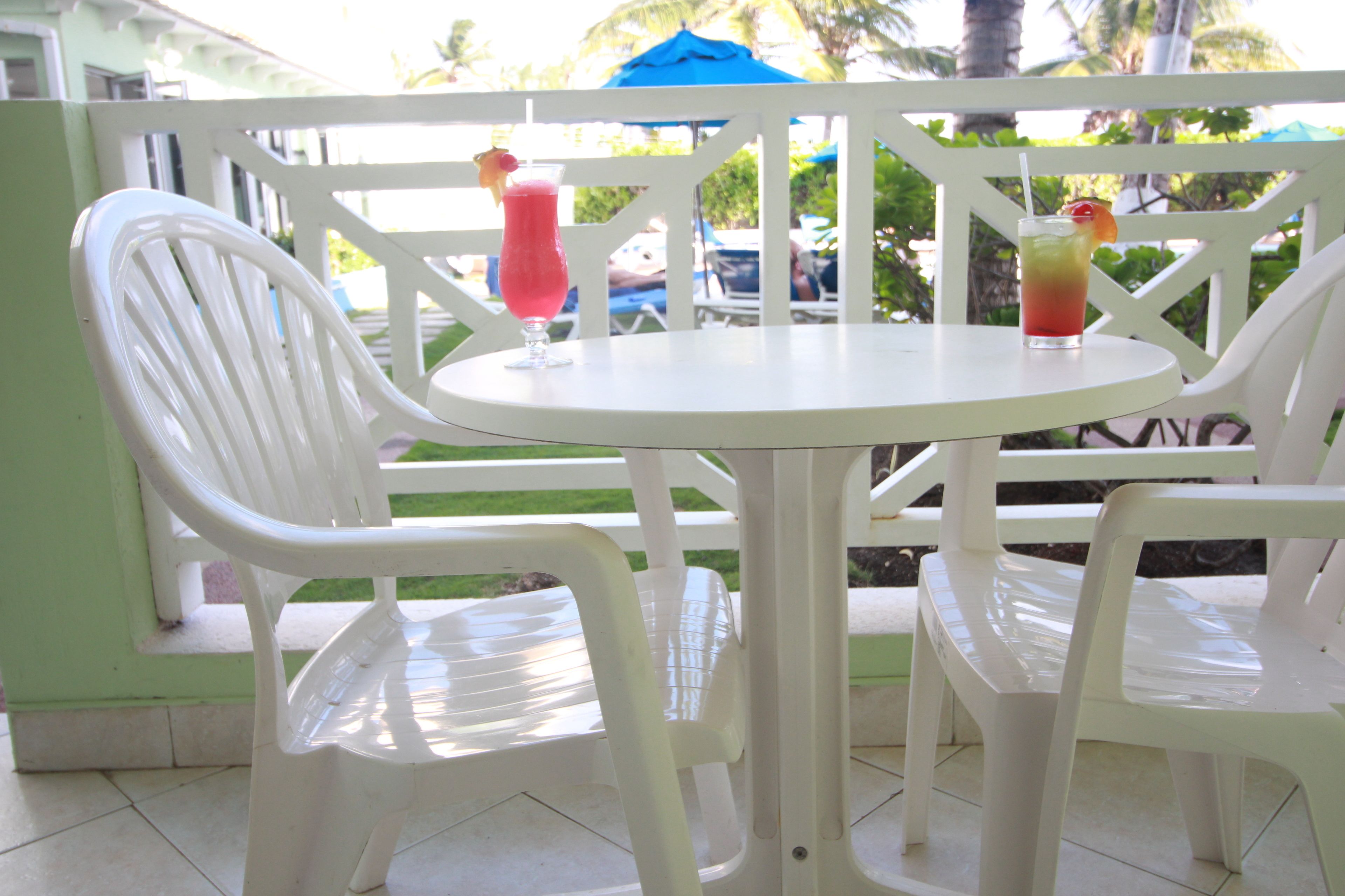 A white plastic table and chairs set on a balcony with a view of palm trees and a blue umbrella.