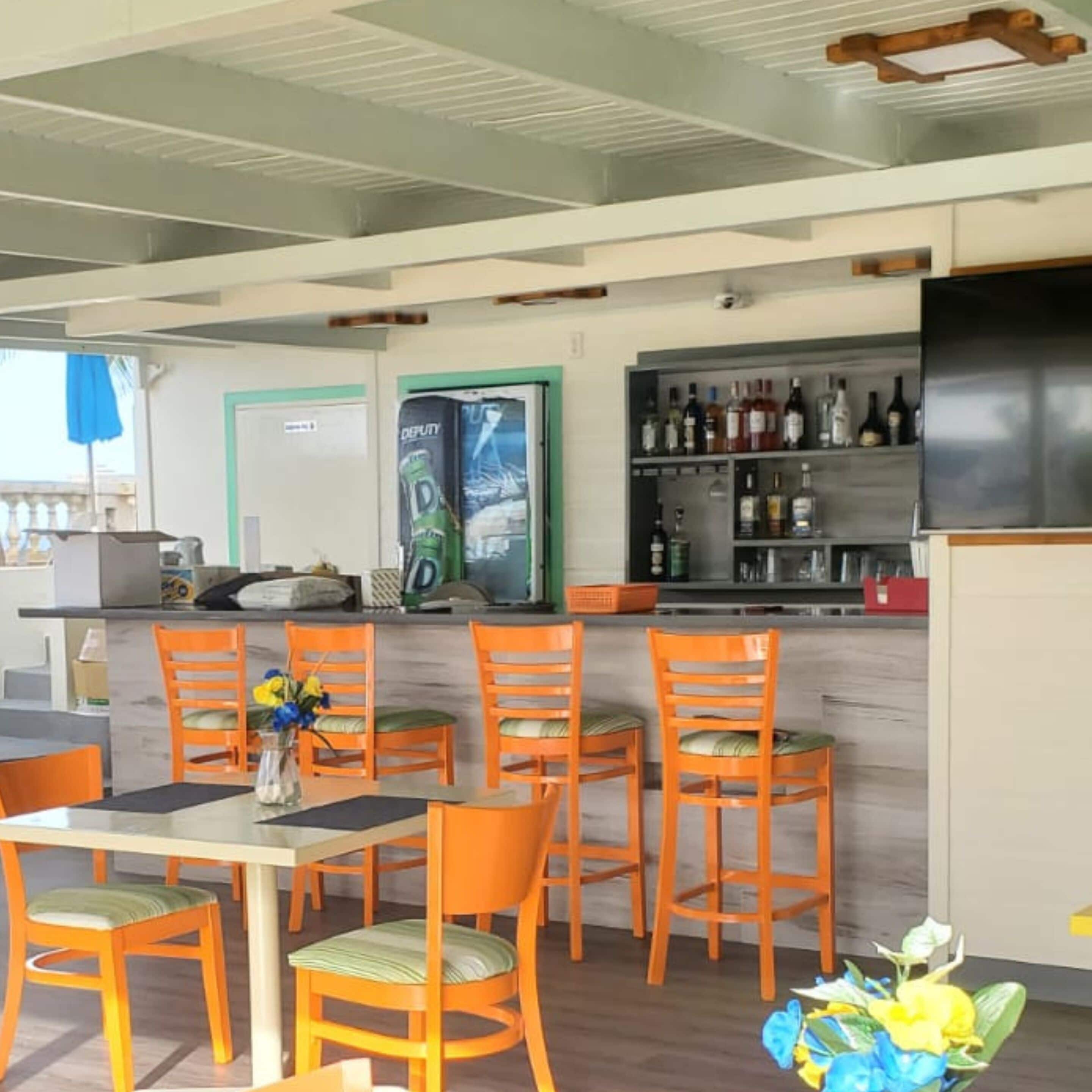 A restaurant with a bar area, multiple orange chairs, a table with a vase of flowers, and a shelf with bottles.