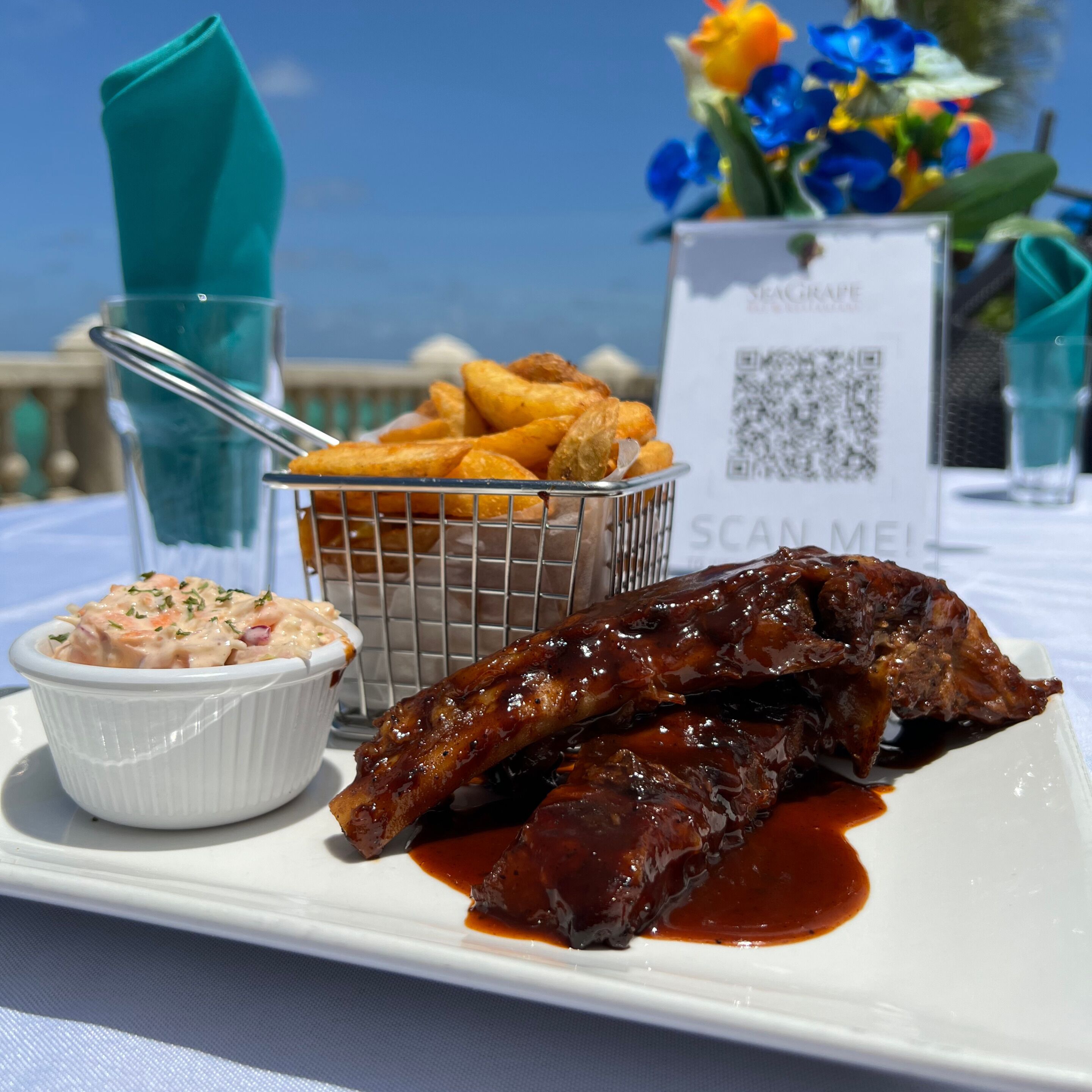 A plate with ribs and a side of coleslaw, a basket of fries, and a QR code on a table.