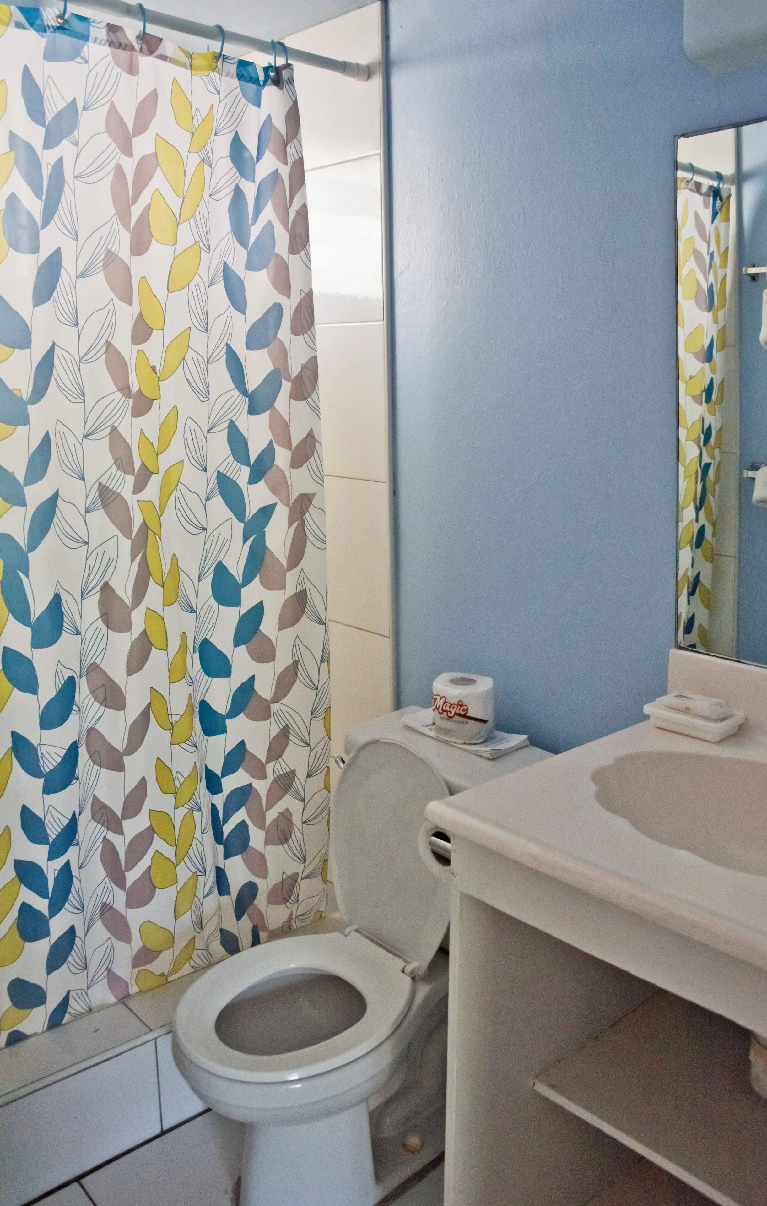 Un baño con inodoro, lavamanos, espejo y cortina de ducha estampada con hojas.