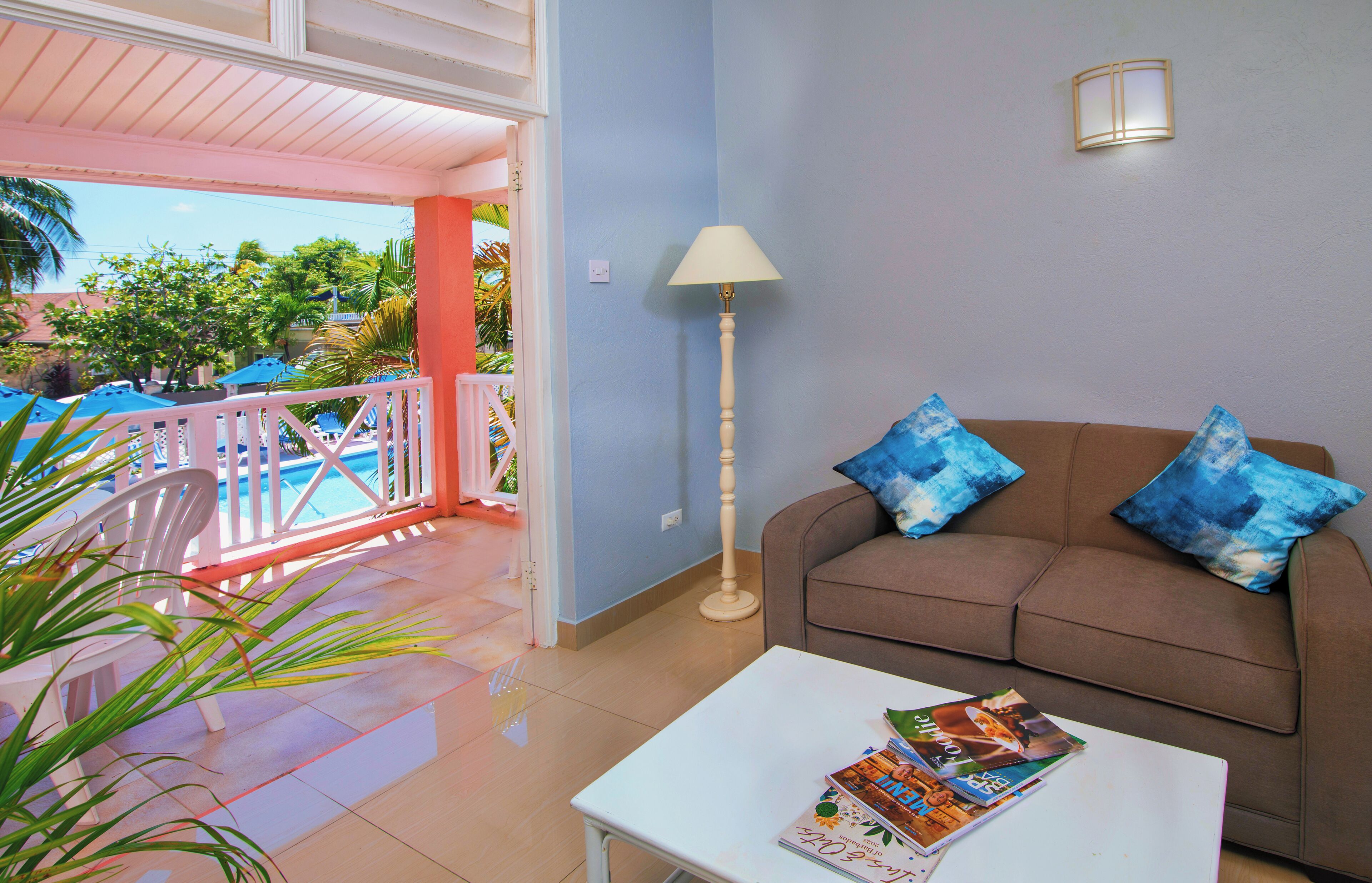 A living room with a brown sofa, two blue pillows, a white coffee table with magazines, a floor lamp, and a view of a pool area with a pink railing.