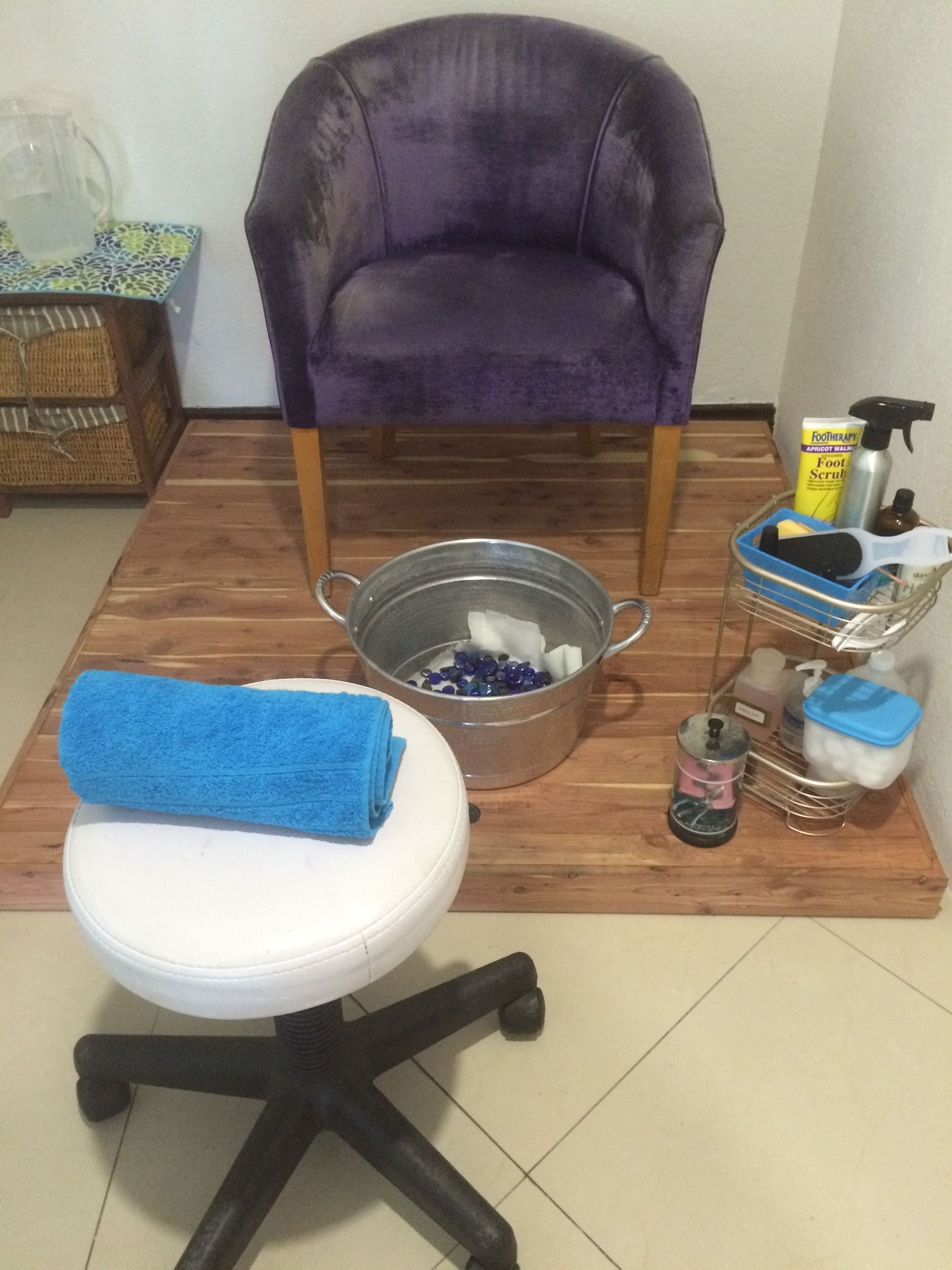 Quarto com uma cadeira roxa, um banquinho branco, uma bacia de metal com contas, uma toalha azul e diversos produtos de higiene pessoal sobre um piso de madeira.