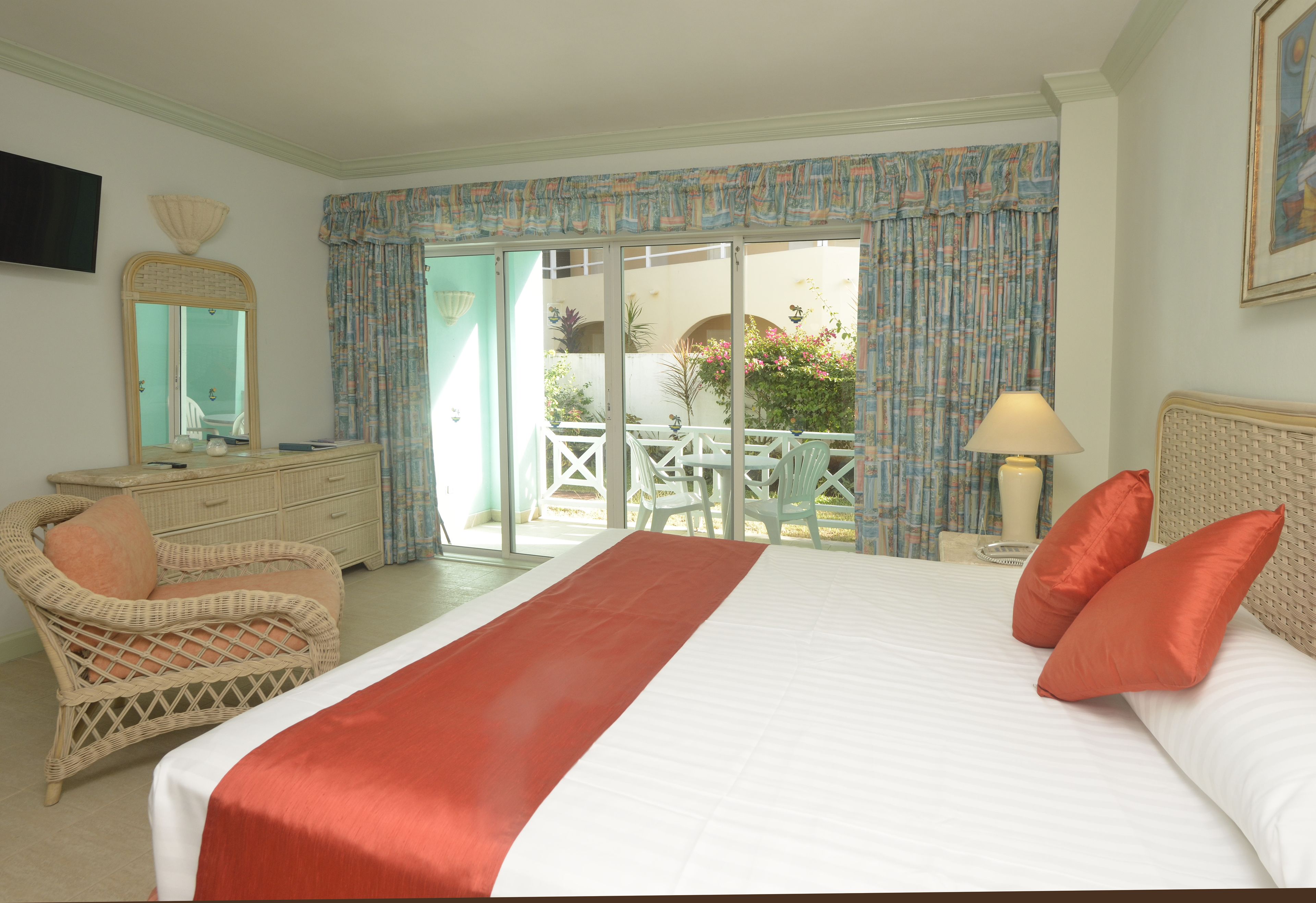 A hotel room with a large bed, a wicker chair, a desk, and a balcony with a table and chairs.