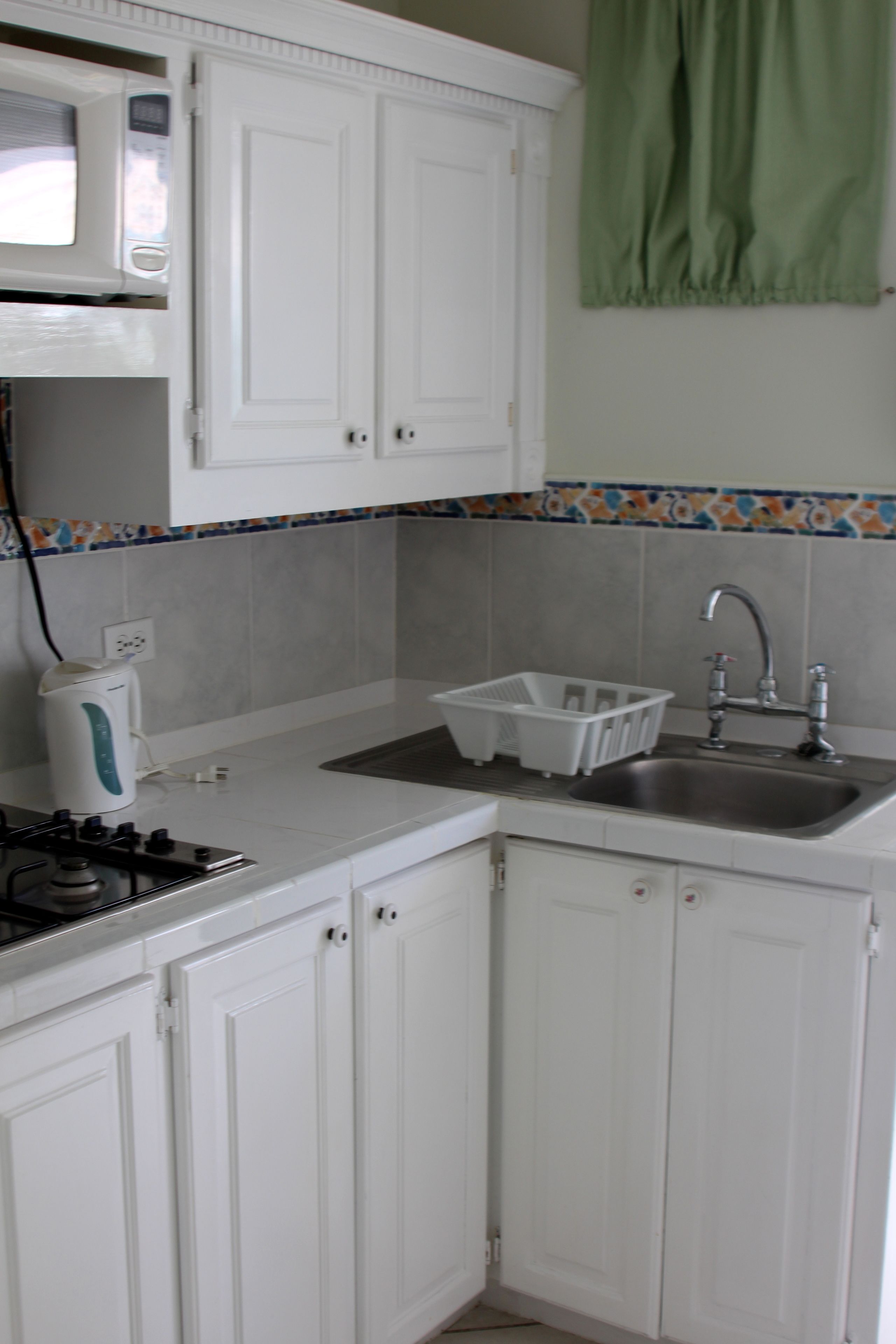 A kitchen with white cabinets, a microwave, a kettle, a sink, and a green curtain.