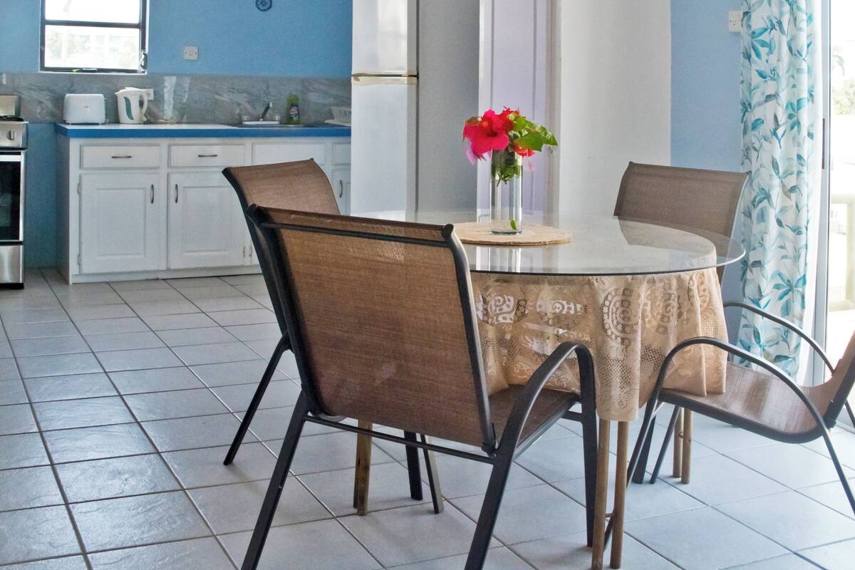 Un comedor con una mesa redonda, cuatro sillas, un jarrón con flores y una cocina al fondo.