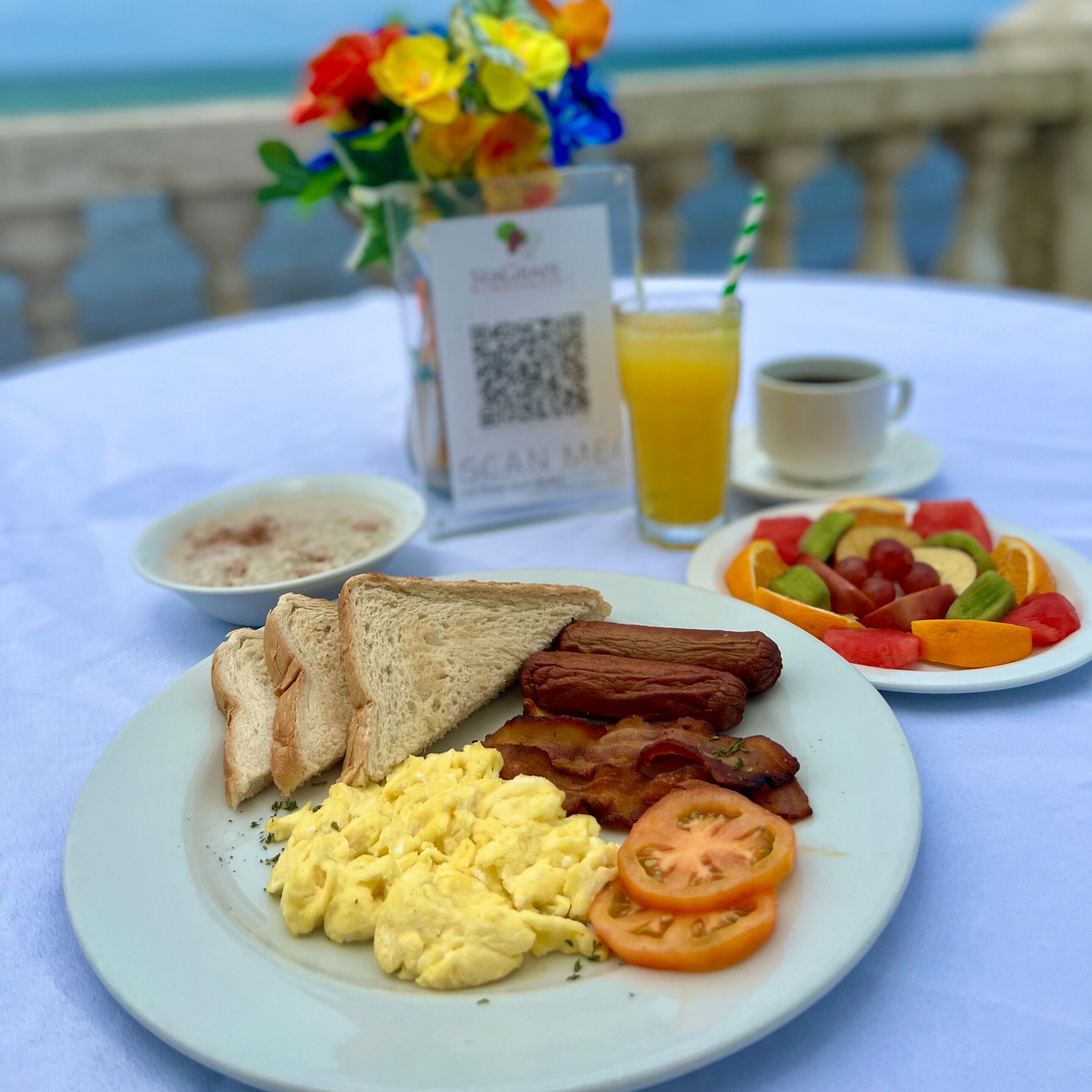 A breakfast plate with toast, bacon, eggs, and tomatoes, a fruit plate, a glass of orange juice, and a cup of coffee on a table with a QR code and a flower arrangement in the background.