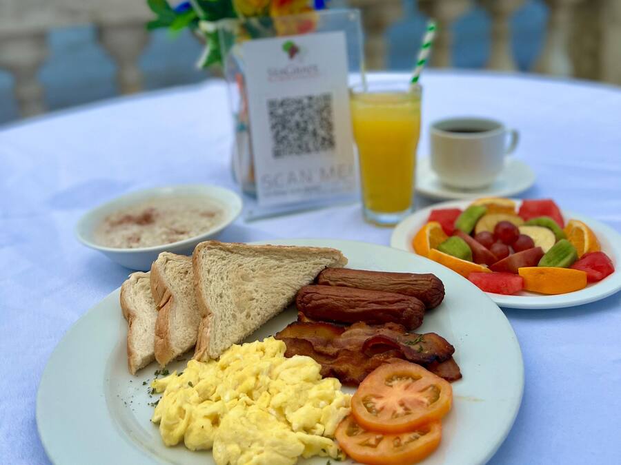Un plato de desayuno con tostadas, tocino, huevos y tomates, un plato de fruta, un vaso de jugo de naranja y una taza de café sobre una mesa con un código QR y un arreglo floral al fondo.