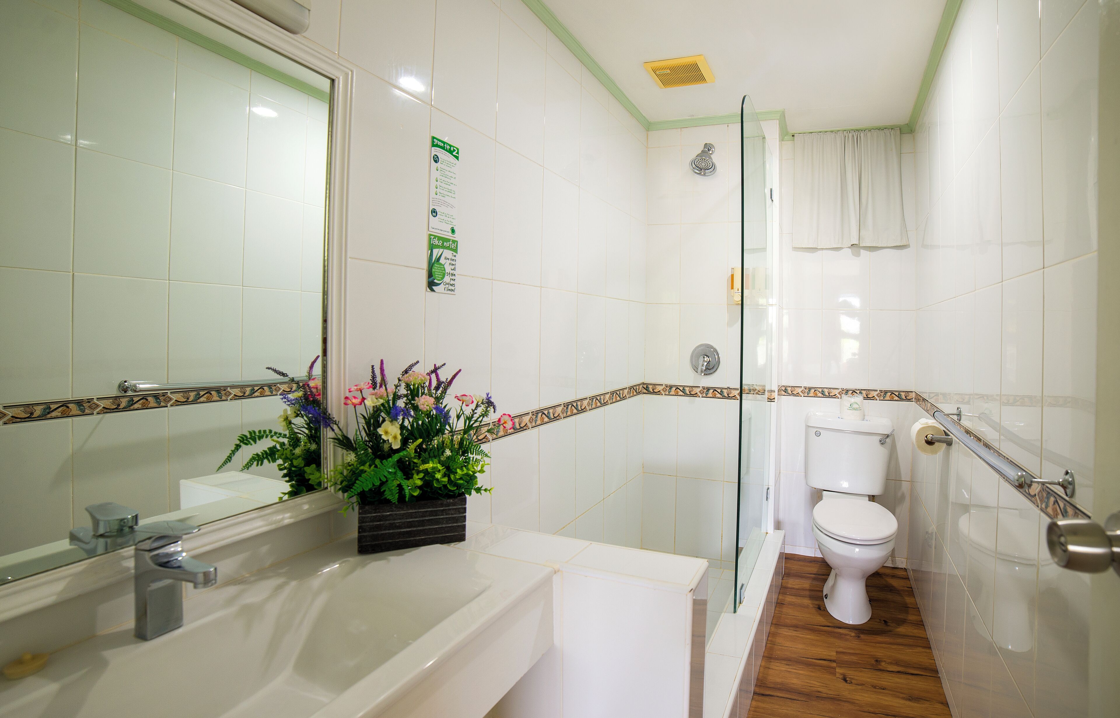 Un baño con lavamanos blanco, inodoro y ducha. Hay un espejo sobre el lavamanos y una planta en la encimera.