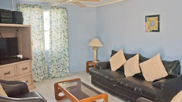 Un salón con sofá, mesa de centro de vidrio, televisor, lámpara y cortinas.