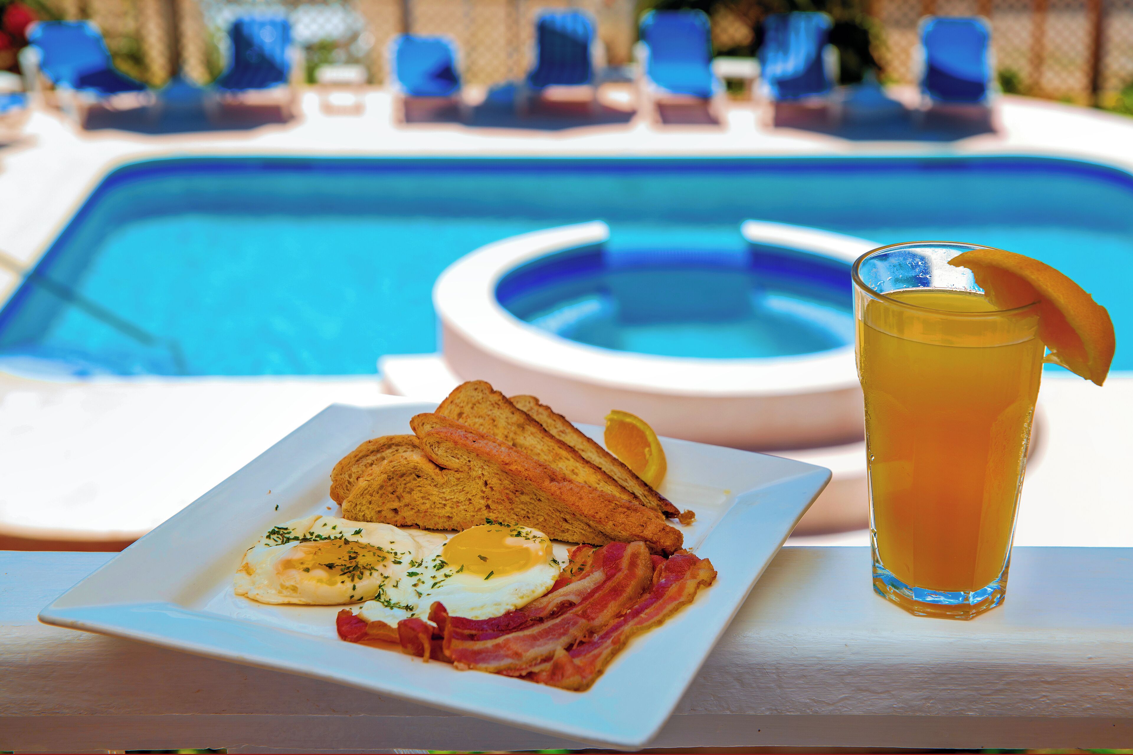 A breakfast plate with eggs, bacon, toast, and a glass of orange juice by a pool.