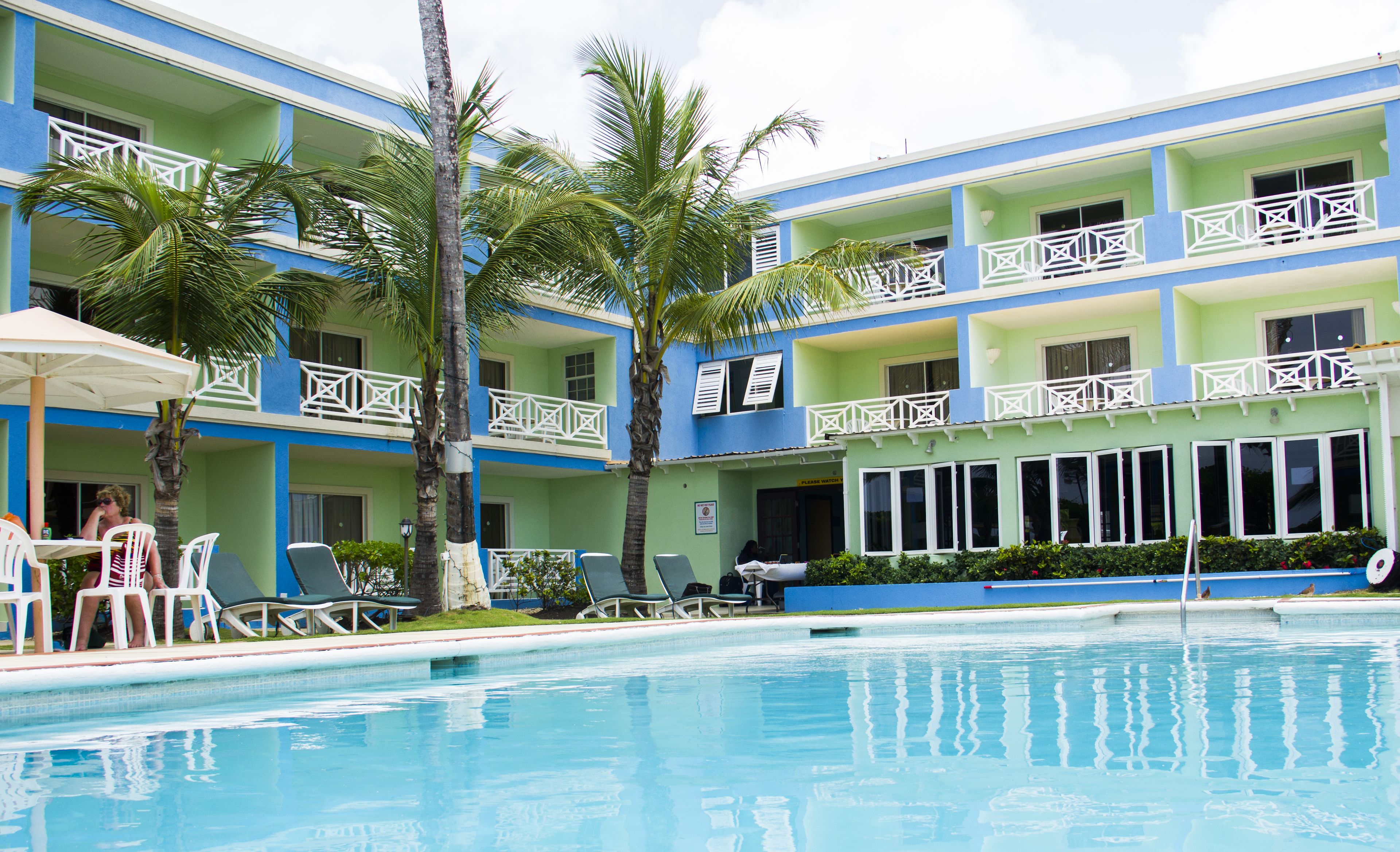Un hotel con zona de piscina, que cuenta con varios balcones con barandillas blancas, palmeras y sillones de descanso.
