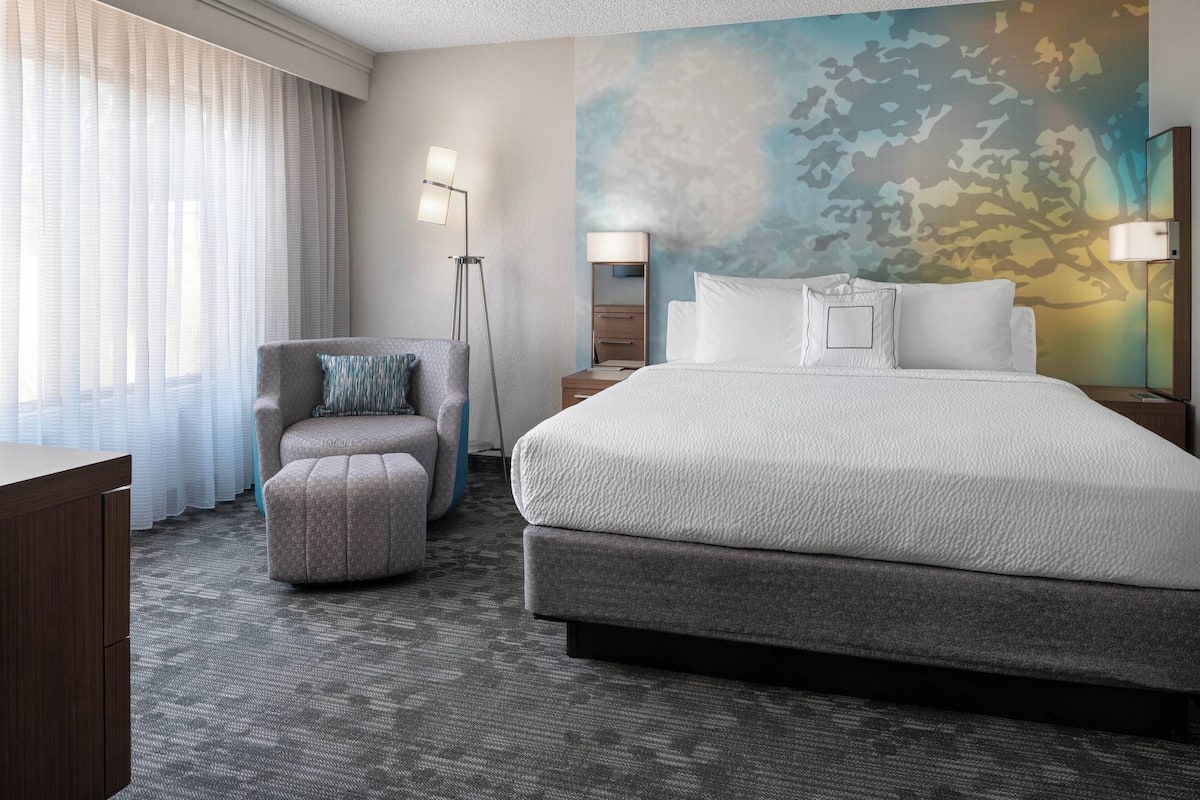 Quarto de hotel com uma cama grande, uma cadeira, uma escrivaninha e um abajur.