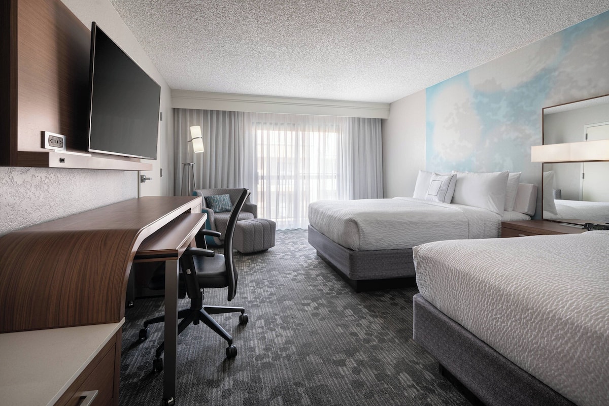 Quarto de hotel com televisão de tela plana, escrivaninha com cadeira, duas camas, mesa de cabeceira e uma janela grande com cortinas.