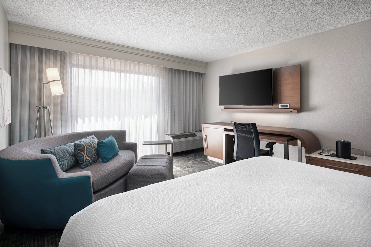 Quarto de hotel moderno com uma cama grande, uma escrivaninha, TV de tela plana e um sofá com almofadas decorativas.