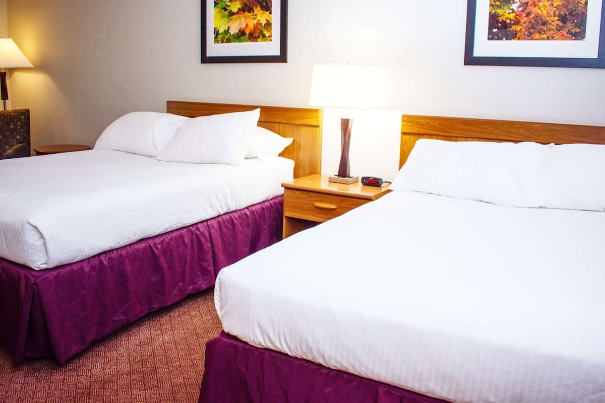 Two adjacent hotel beds with white linens, a wooden headboard, a nightstand with a lamp, and a framed picture on the wall.