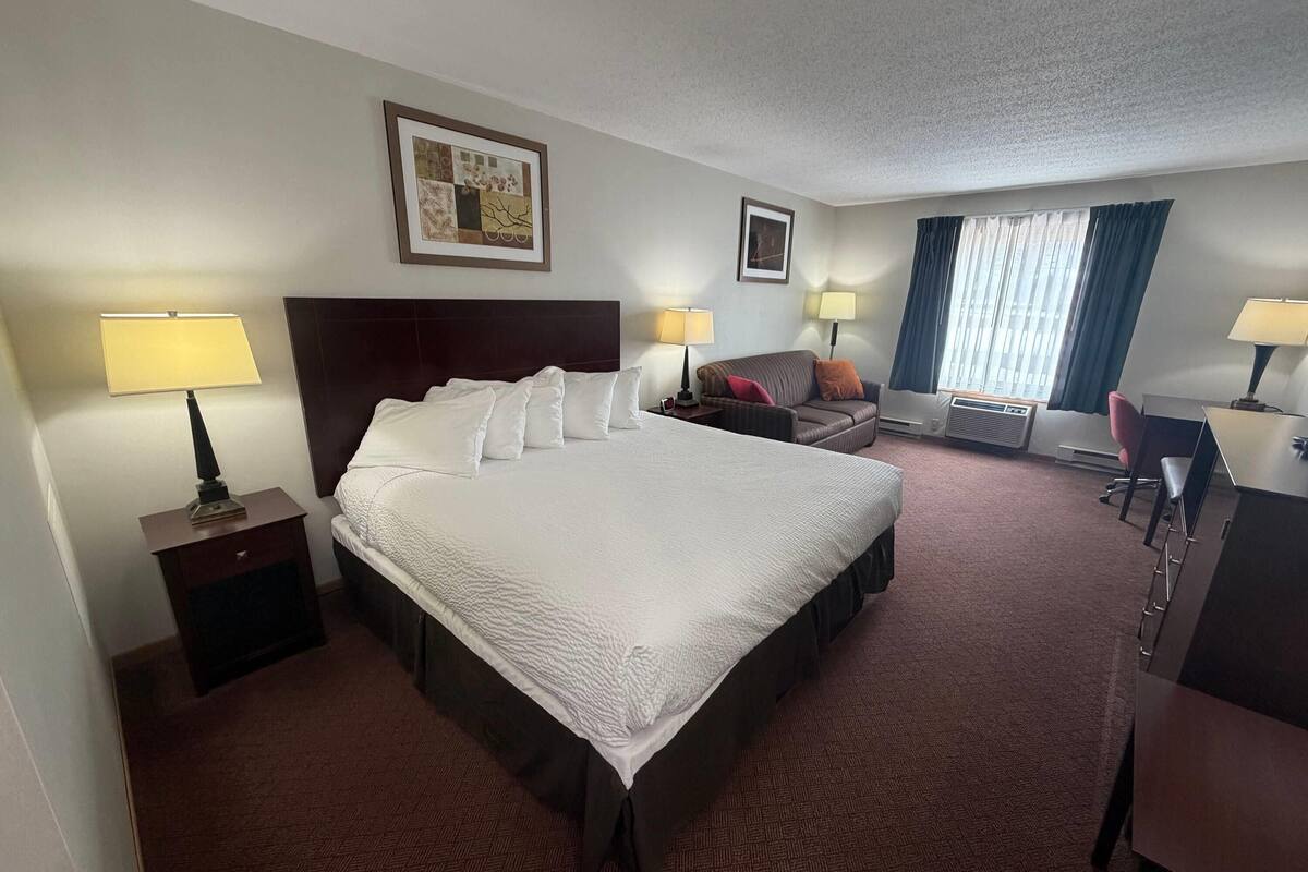 A hotel room with a large bed, two bedside tables with lamps, a desk, a chair, a sofa, and a window with curtains.