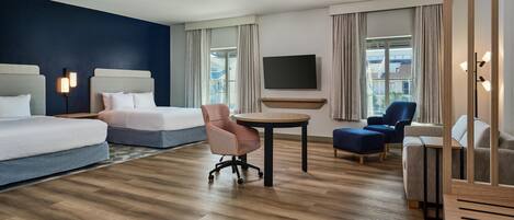 Habitación de hotel con cama, escritorio, silla, televisor y vista a edificios a través de la ventana.