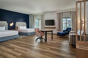Habitación de hotel con cama, escritorio, silla, televisor y vista a edificios a través de la ventana.