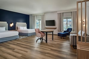 Quarto de hotel com cama, escrivaninha, cadeira, televisão e vista para edifícios pela janela.