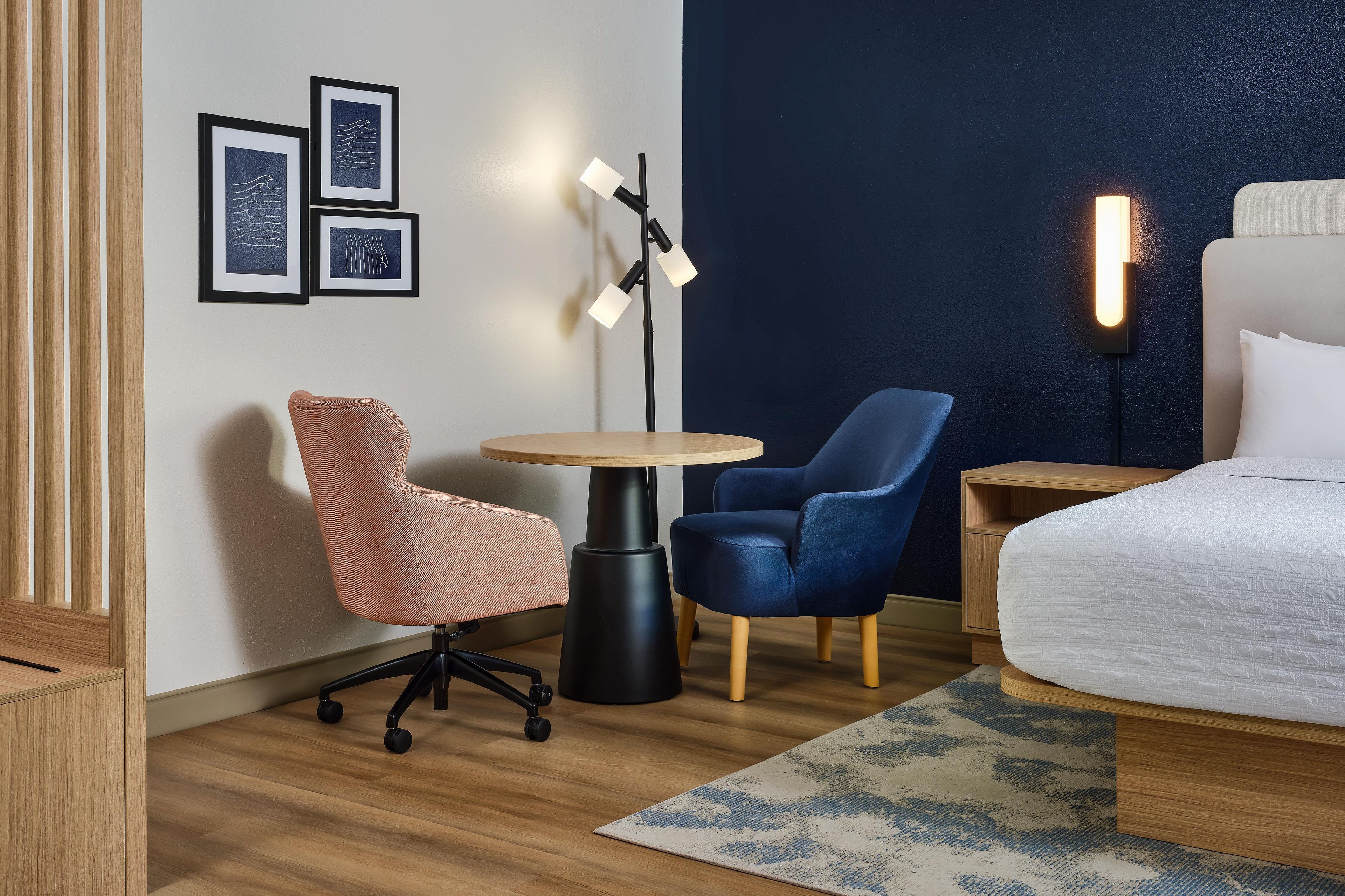 Habitación de hotel con cama, dos sillones, una mesita, l ámpara de pie y cuadros en la pared.