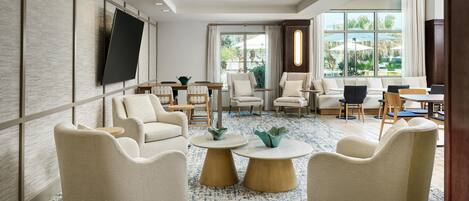 A modern hotel lobby with a large TV, comfortable seating, a coffee table, and a view of the outdoors.