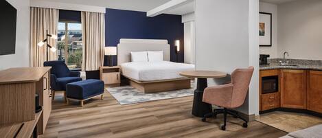 Habitación de hotel con cama, escritorio, silla, kitchenette y vistas al exterior.