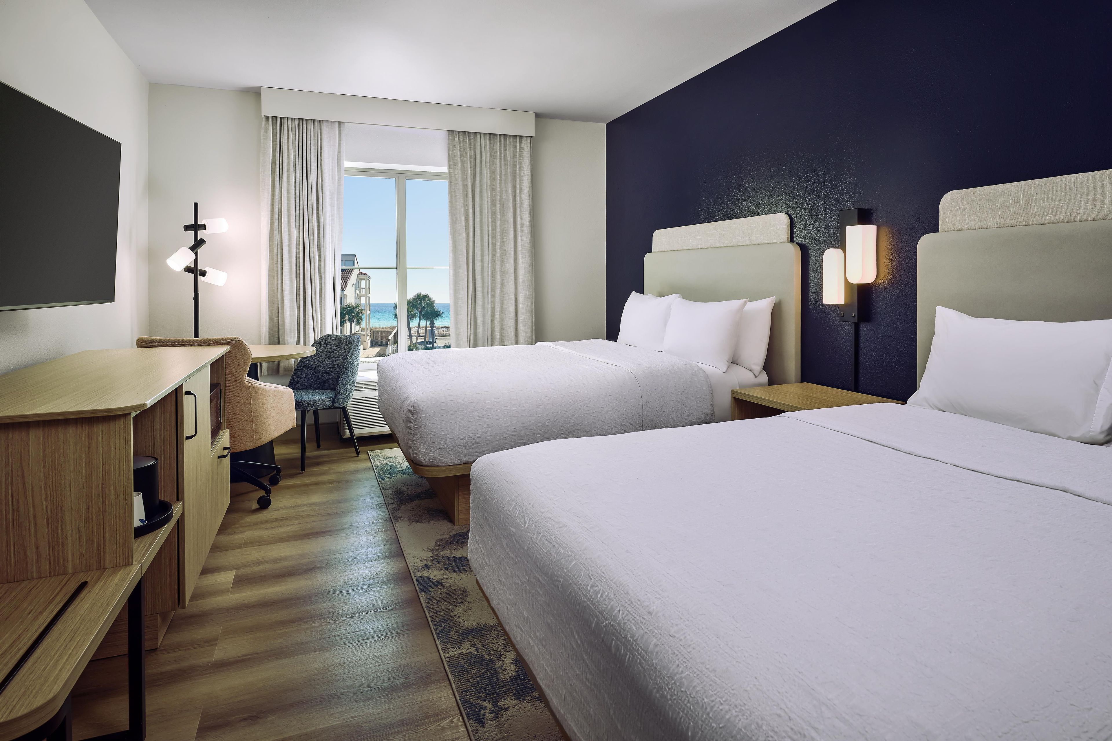 A hotel room with two beds, a desk, a chair, a TV, and a view of the beach.