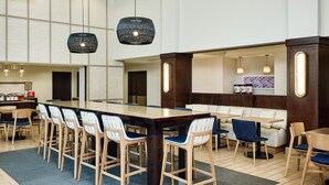 Un moderno vestíbulo de hotel con una mesa común alargada, taburetes de bar, un sofá y una zona de cocina visible a través de un panel de vidrio.
