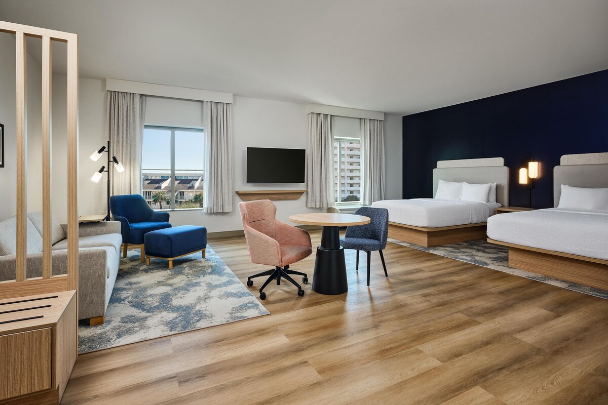 A hotel room with a bed, a desk, two chairs, a TV, and a view of a building.