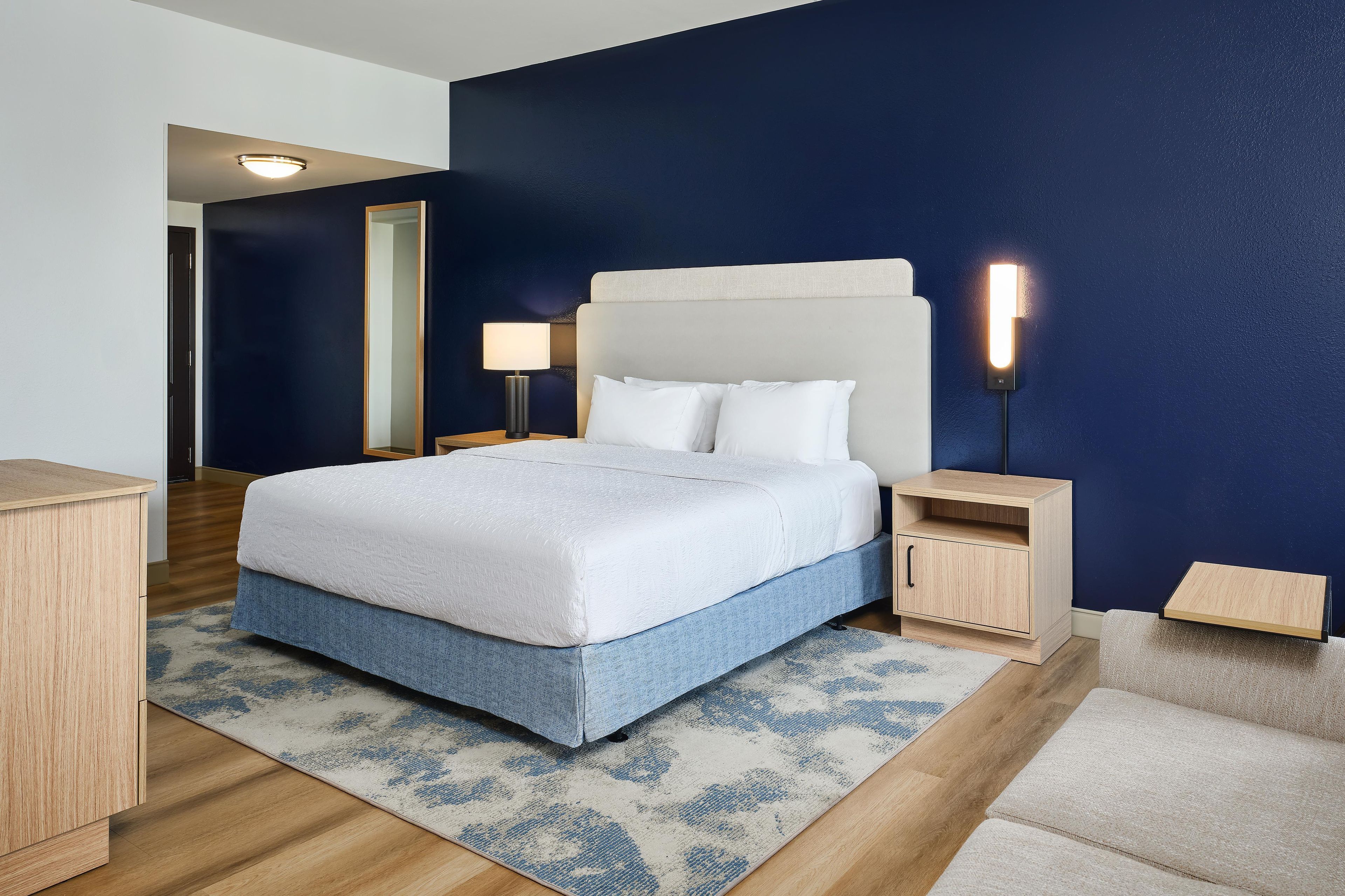 Quarto de hotel moderno com uma cama grande, mesas de cabeceira, um criado-mudo e uma luminária na parede.