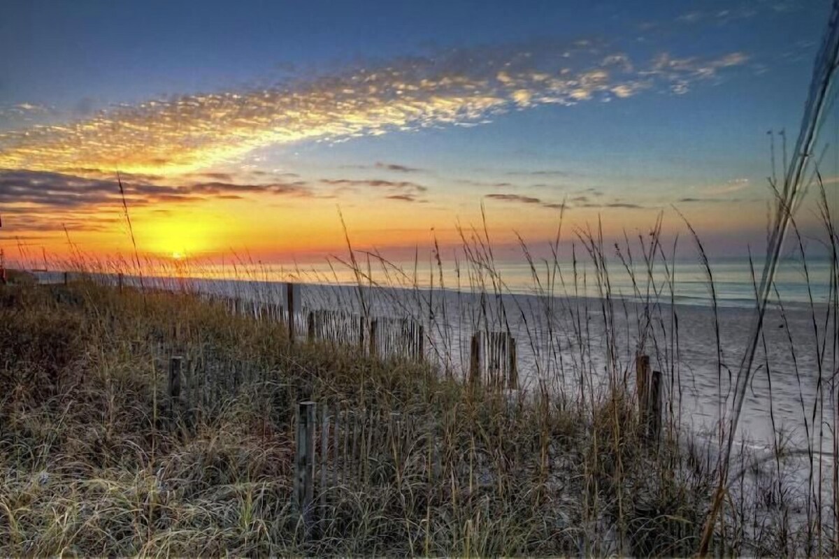 A beach with a sunset, grassy dunes, and a wooden fence.