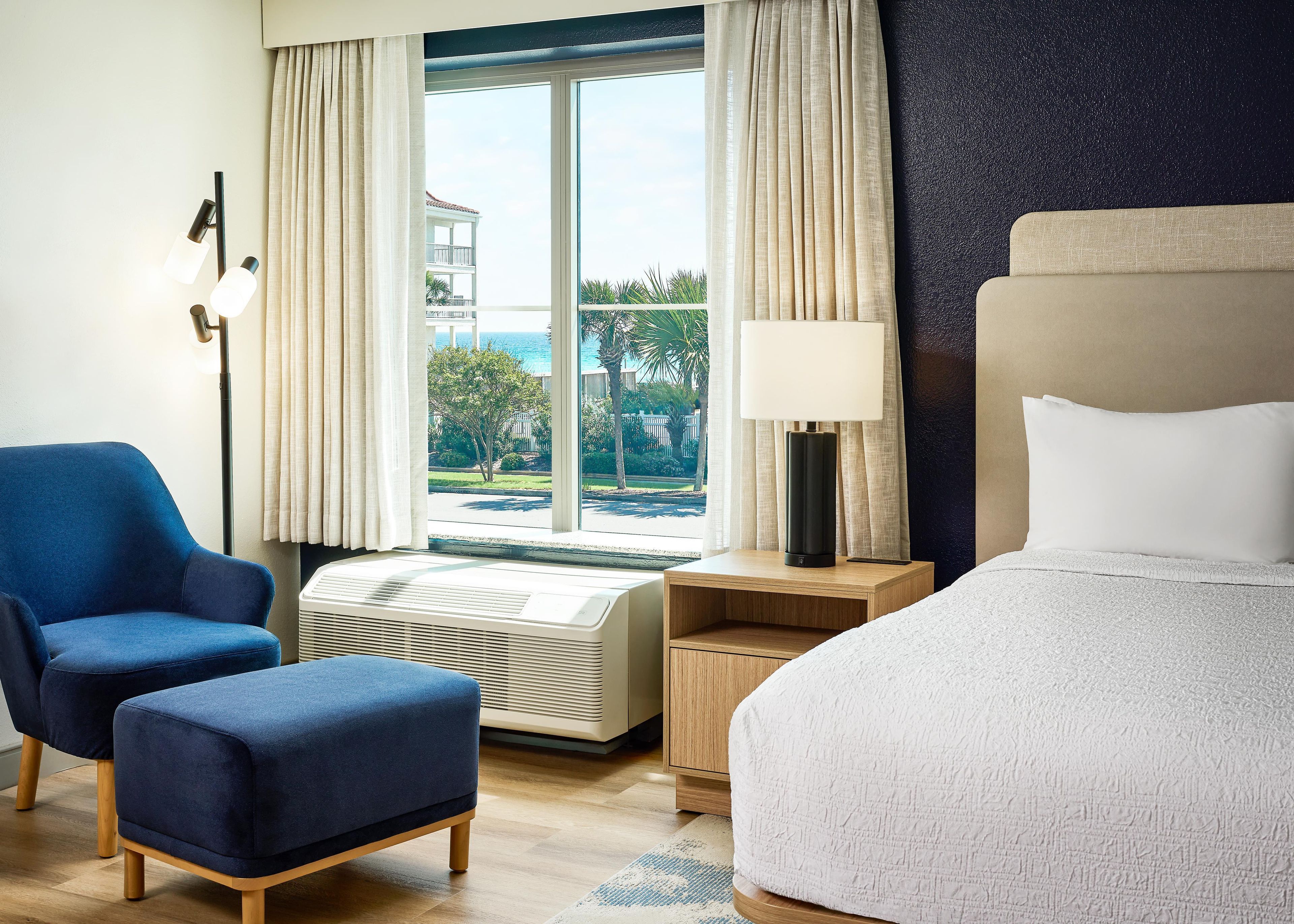 A hotel room with a bed, a blue armchair, a small table with a lamp, a window with curtains, and a view of the outdoors.
