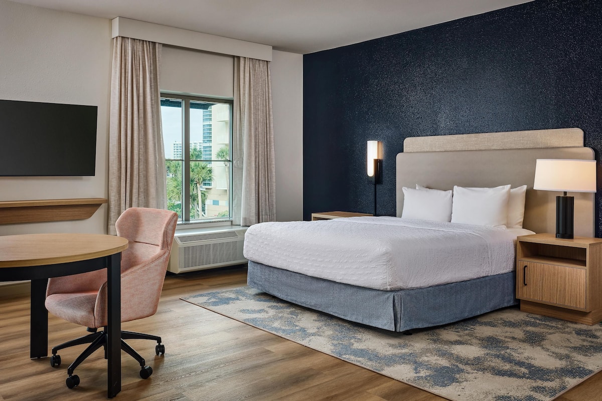 A hotel room with a bed, a desk, a chair, a nightstand, and a window with curtains.