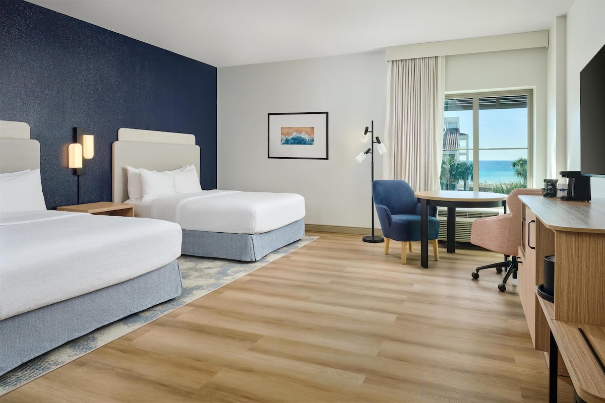 A hotel room with two beds, a desk, a chair, and a large window with a view of the ocean.