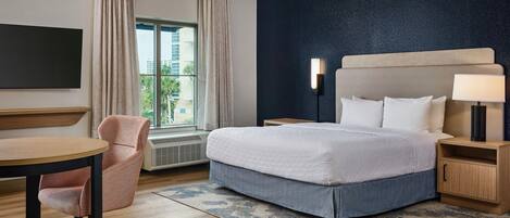 Habitación de hotel con cama, mesita de noche, lámpara, silla y vistas al exterior.
